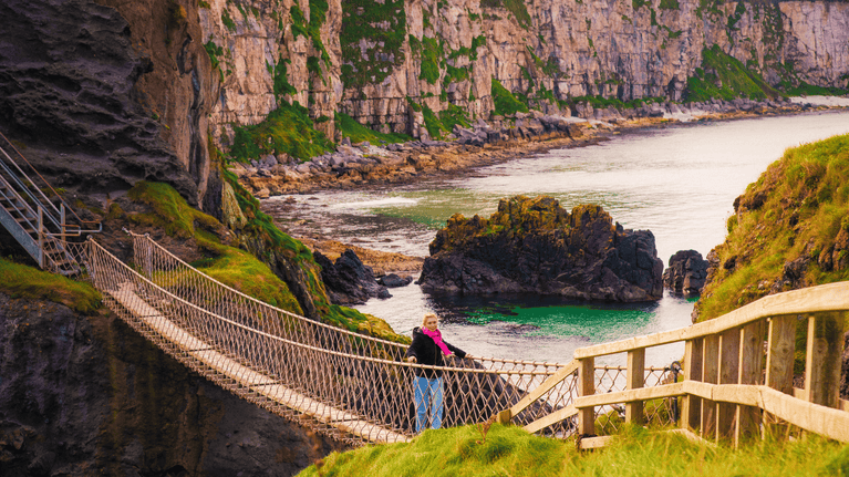 Visitor crossing Carrick-a-Rede Rope Bridge on an autumn day, with rugged coastal cliffs, rocky outcrops and calm turquoise waters below